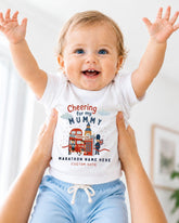 A smiling baby wearing a white t-shirt with a graphic design and text, being held up by someone's arms.