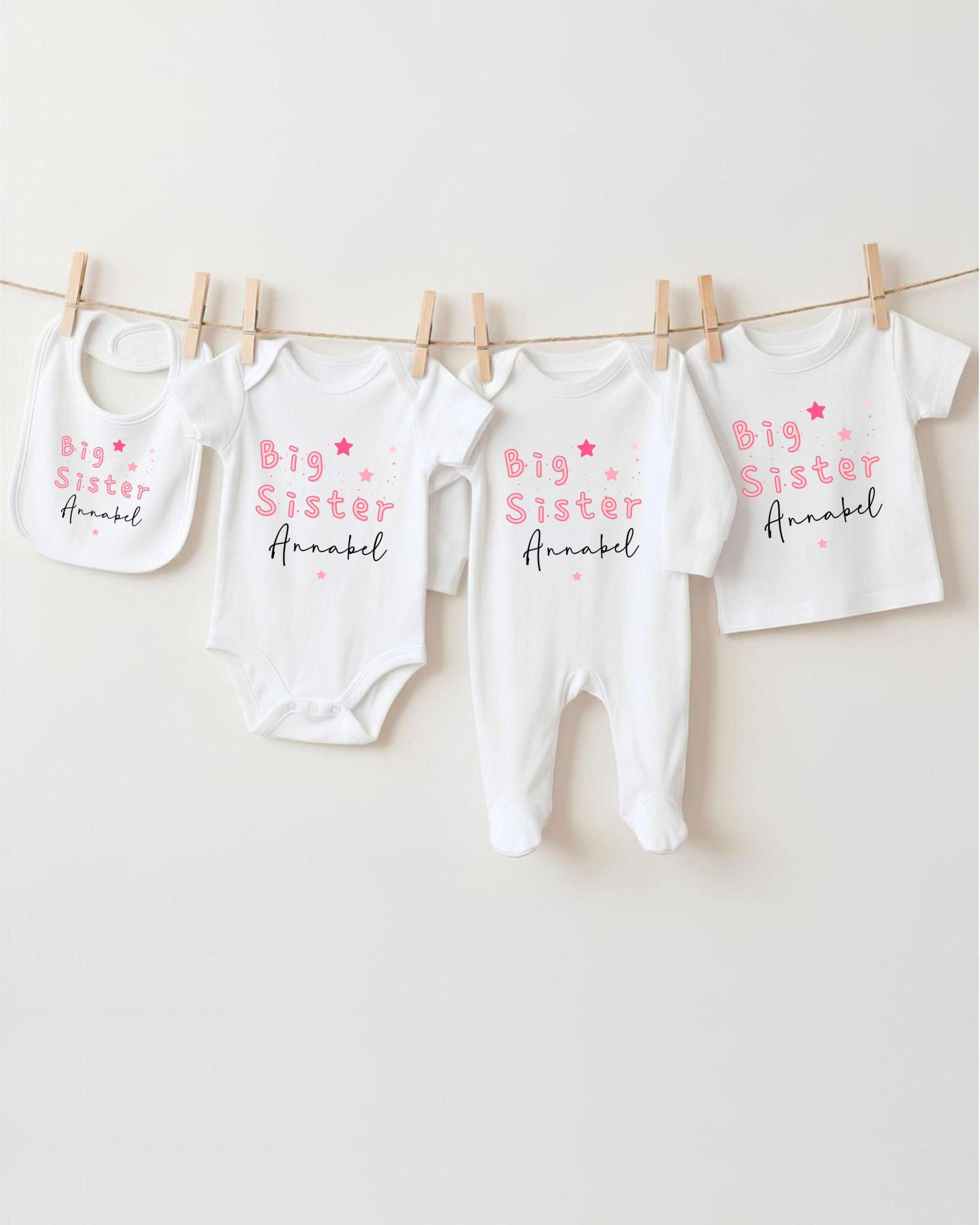A row of four white baby clothes with the text "Big Sister" printed on them, hanging on a clothesline.