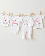 A row of four white baby clothes with the text "Big Sister" printed on them, hanging on a clothesline.