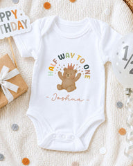 A white onesie with a teddy bear and the text "Half Way to One" printed on it, along with the name "Joshua" below the bear. The onesie is placed on a white blanket, surrounded by birthday decorations such as a "Happy Birthday" sign, a gift box, and confetti.