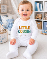 A smiling baby boy wearing a white onesie with the text "LITTLE COUSIN Frankie" is sitting on the floor in a nursery setting.
