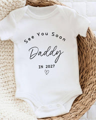 A white baby onesie with the text "See You Soon Daddy in 2027" printed on it.