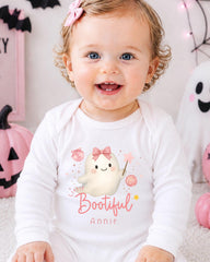 A smiling baby girl wearing a white onesie with a ghost character and the text "Little Bootful Annie" is sitting in front of Halloween decorations.