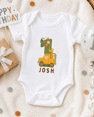 A white onesie with a cartoon image of a giraffe and a monkey, along with the name " Josh" printed on it, is displayed on a bed with a "Happy Birthday" sign and a balloon.
