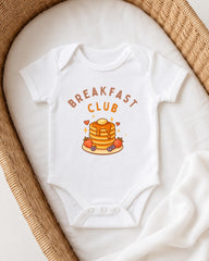 A white onesie with the text "BREAKFAST CLUB" printed on it, featuring a stack of pancakes and various breakfast-themed illustrations.