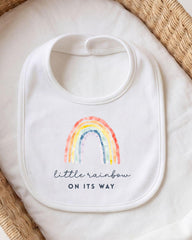 A white bib with a rainbow graphic and the text "little rainbow on its way" printed on it.