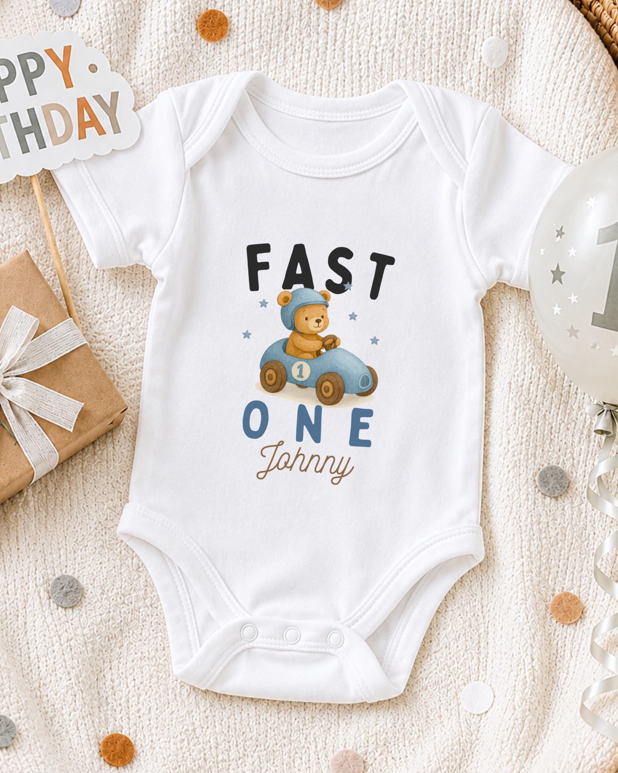 A white onesie with a cartoon image of a bear in a car and the text "FAST ONE Johnny" printed on it, along with a "Happy Birthday" sign and a balloon.