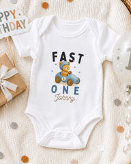 A white onesie with a cartoon image of a bear in a car and the text "FAST ONE Johnny" printed on it, along with a "Happy Birthday" sign and a balloon.