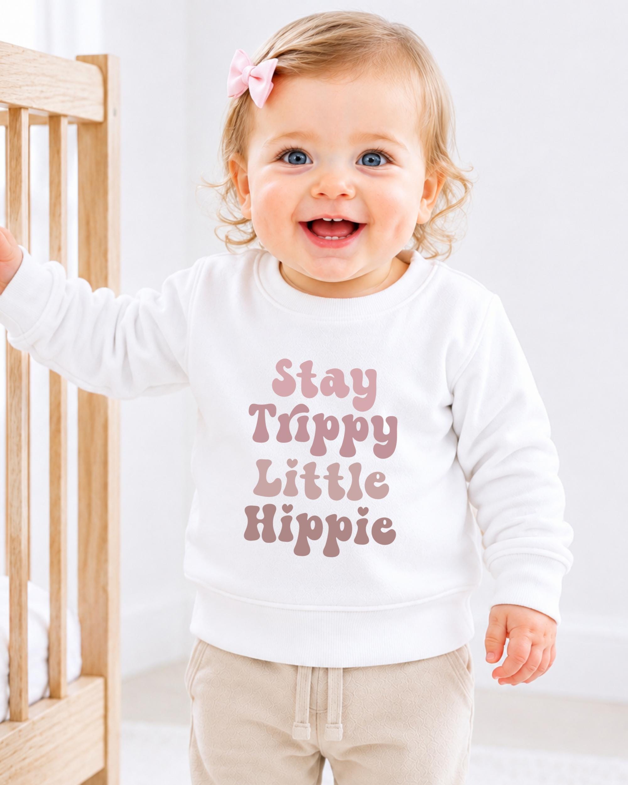 A smiling baby girl wearing a white sweatshirt with the text "Stay Trippy Little Hippie" printed on it.