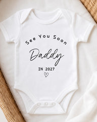 A white baby onesie with the text "See You Soon Daddy in 2027" printed on it.