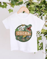 A white t-shirt with a graphic design featuring a dinosaur and the text "OSCAR" on it, hanging on a wooden hanger.