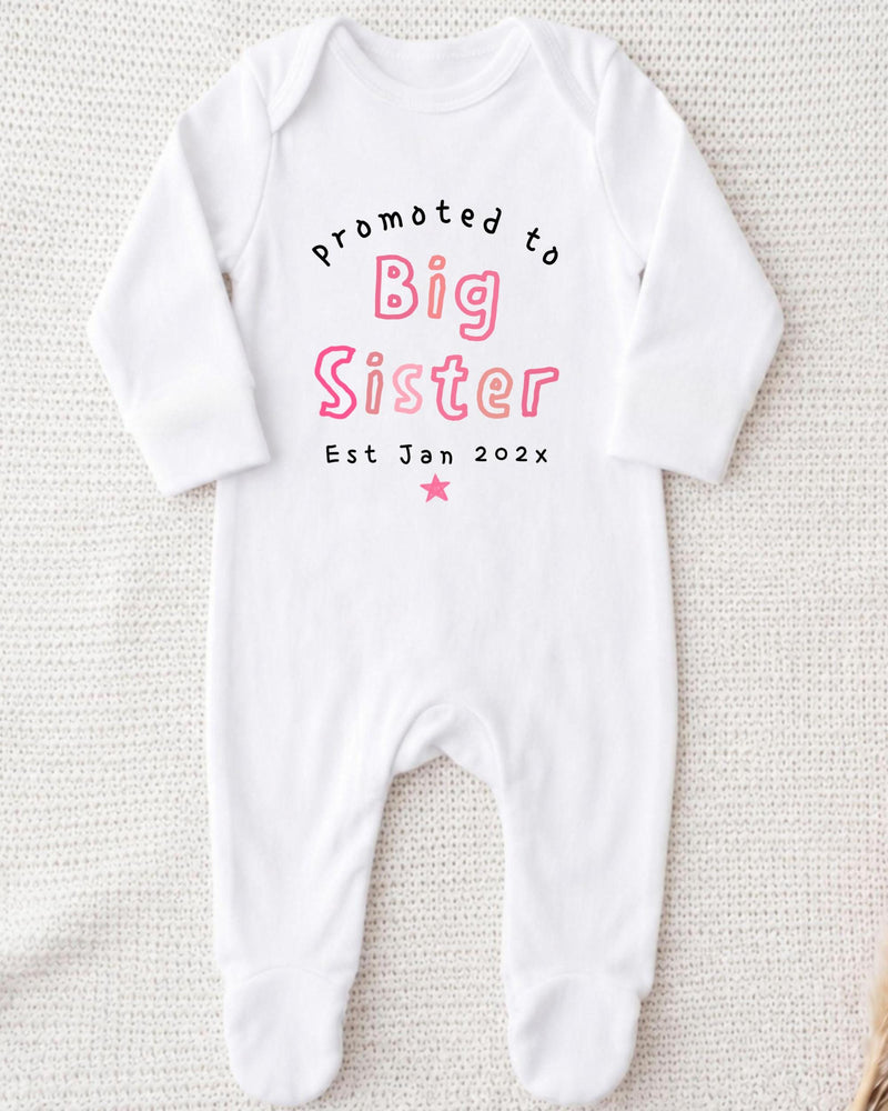 A white bodysuit with text printed on it, which appears to be a promotional item for a baby sister.