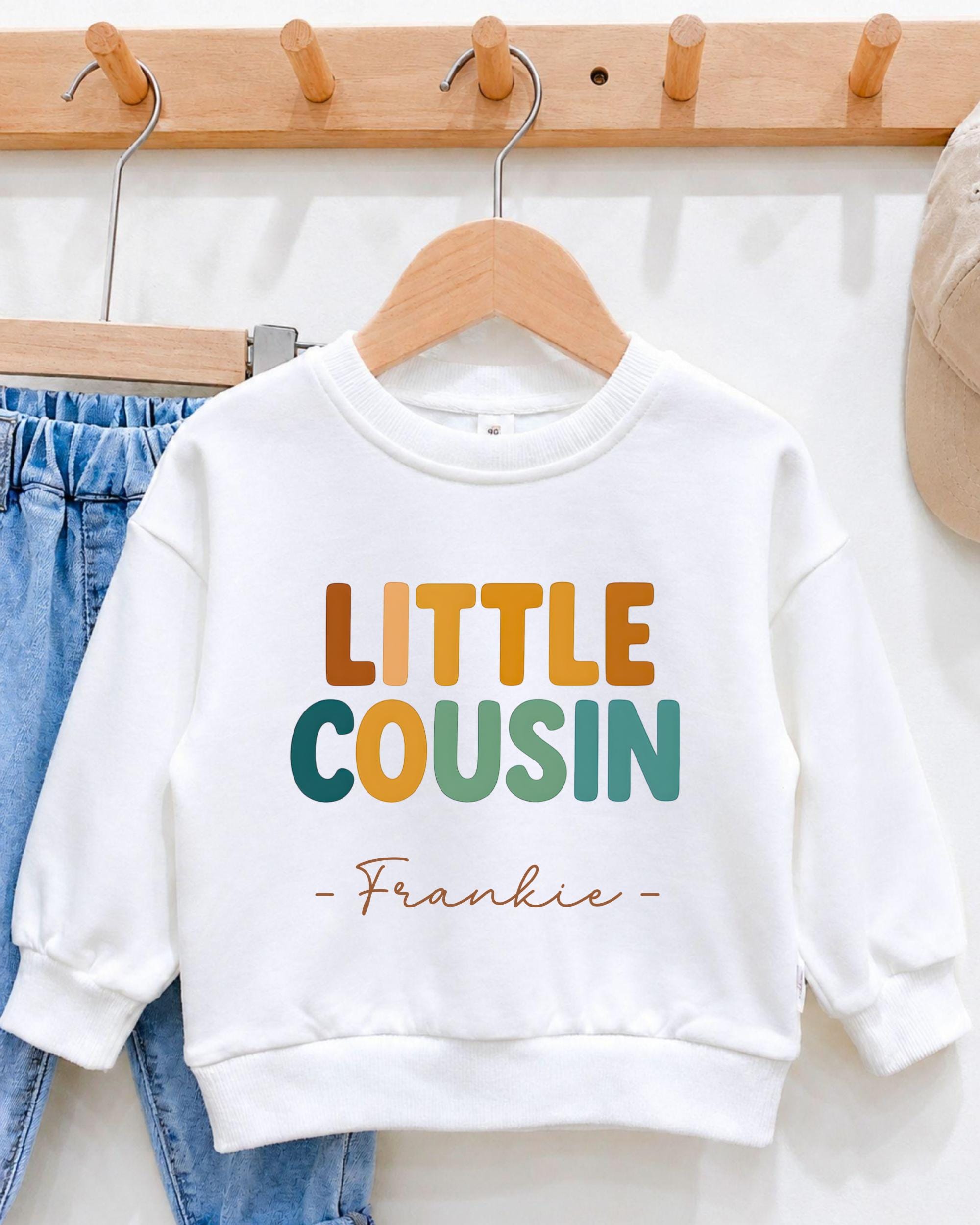 A white sweatshirt with the text "LITTLE COUSIN" printed on it, along with the name "Frankie" below it.