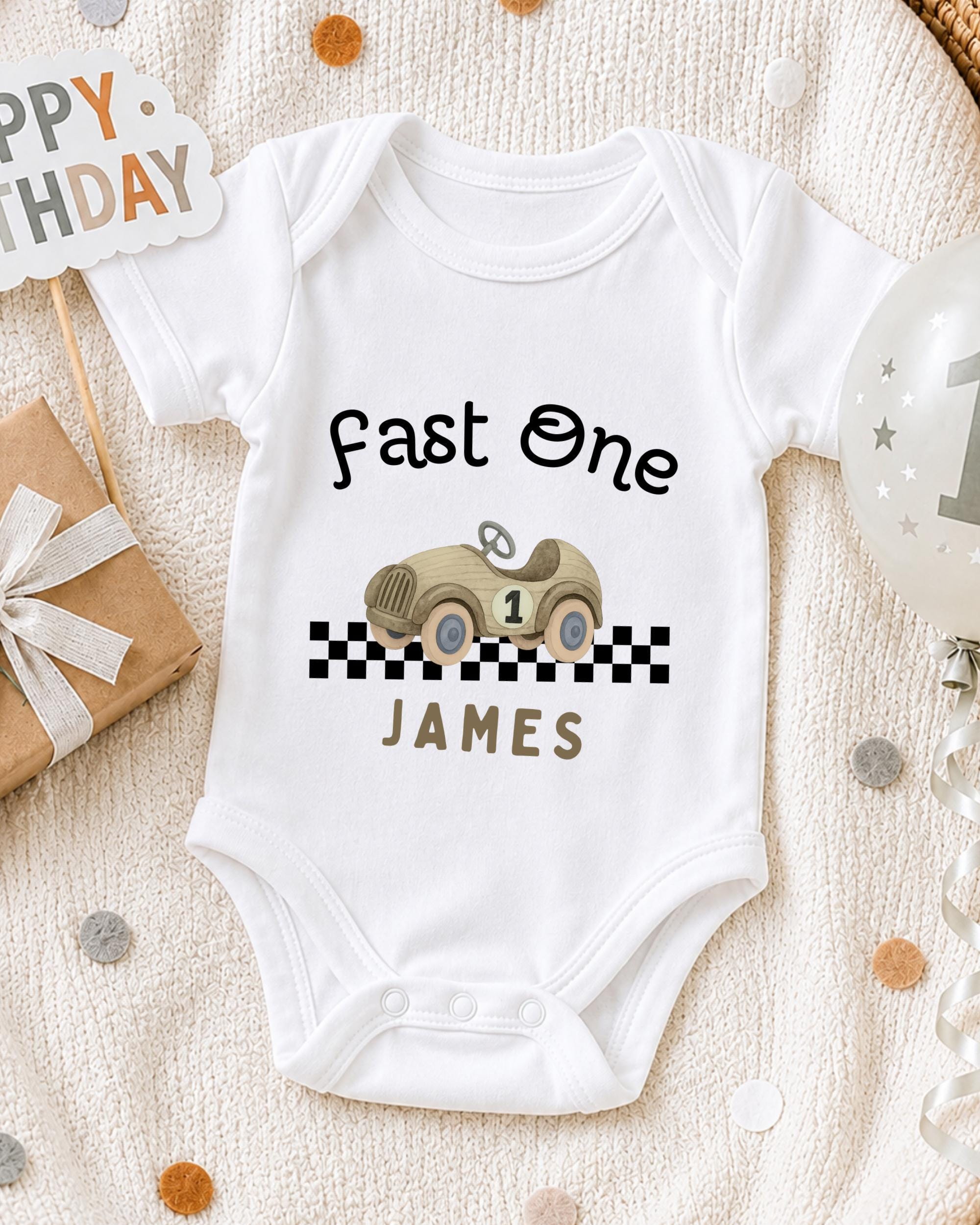 A white onesie with a racing car design and the text "Fast One James" printed on it, along with a birthday sign and a balloon.