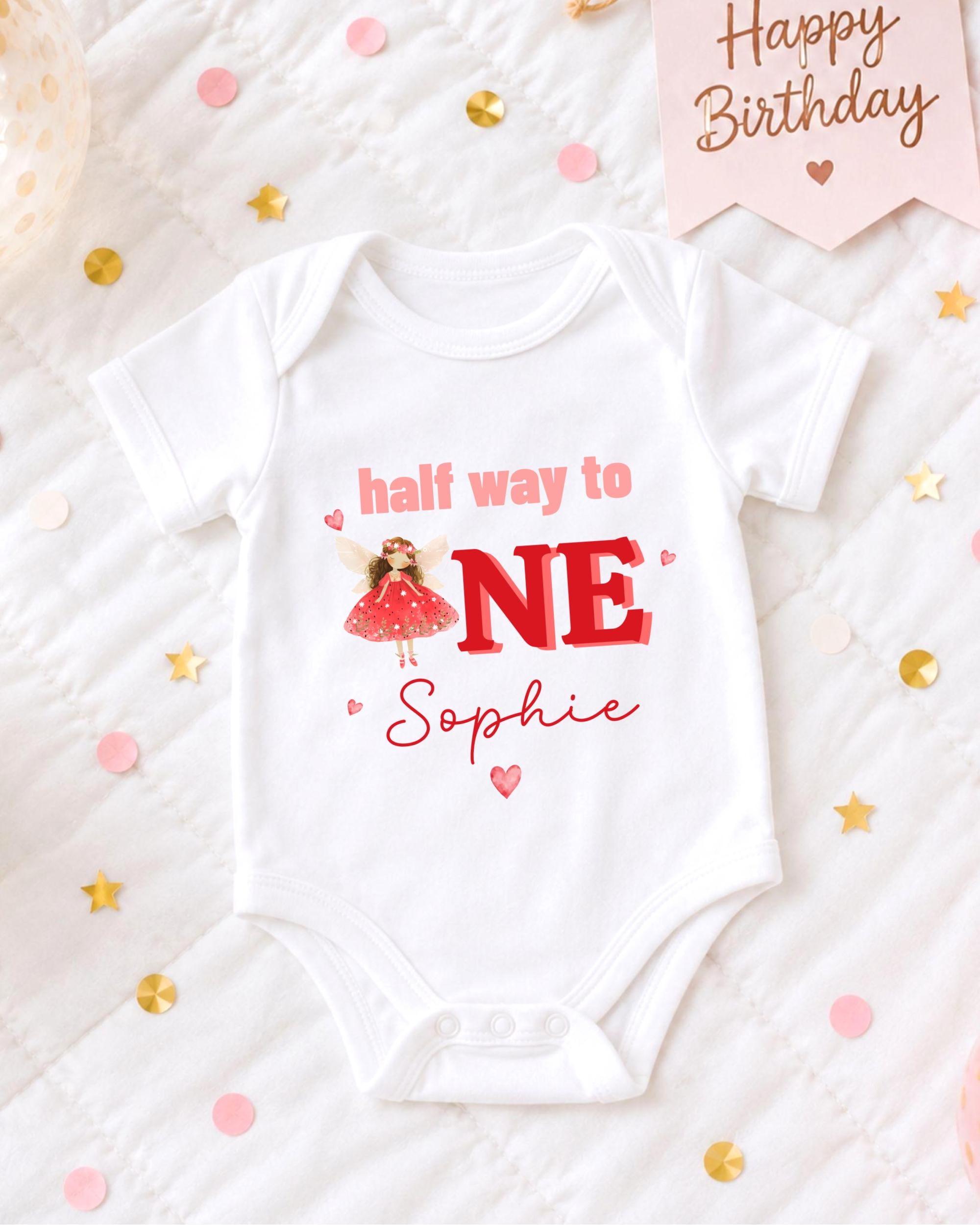 A white onesie with a red design and text, placed on a bed with confetti and a birthday card.