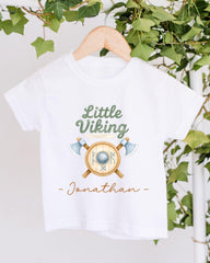 A white t-shirt with a graphic design featuring a shield and crossed axes, along with the text "Little Viking Jonathan" printed on it.