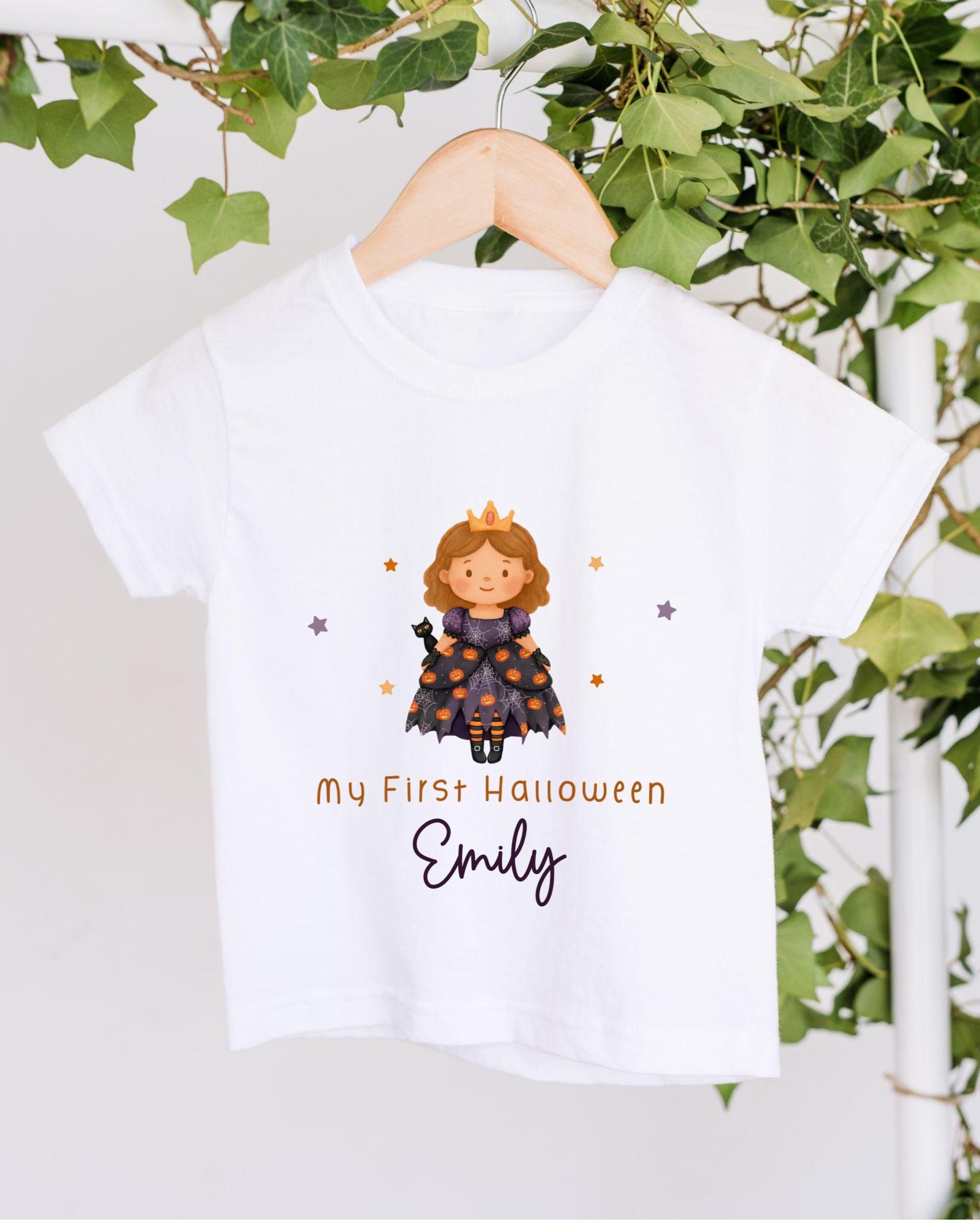 A white t-shirt with a cartoon image of a girl wearing a crown and a purple dress, along with the text "My First Halloween Emily" printed on it.