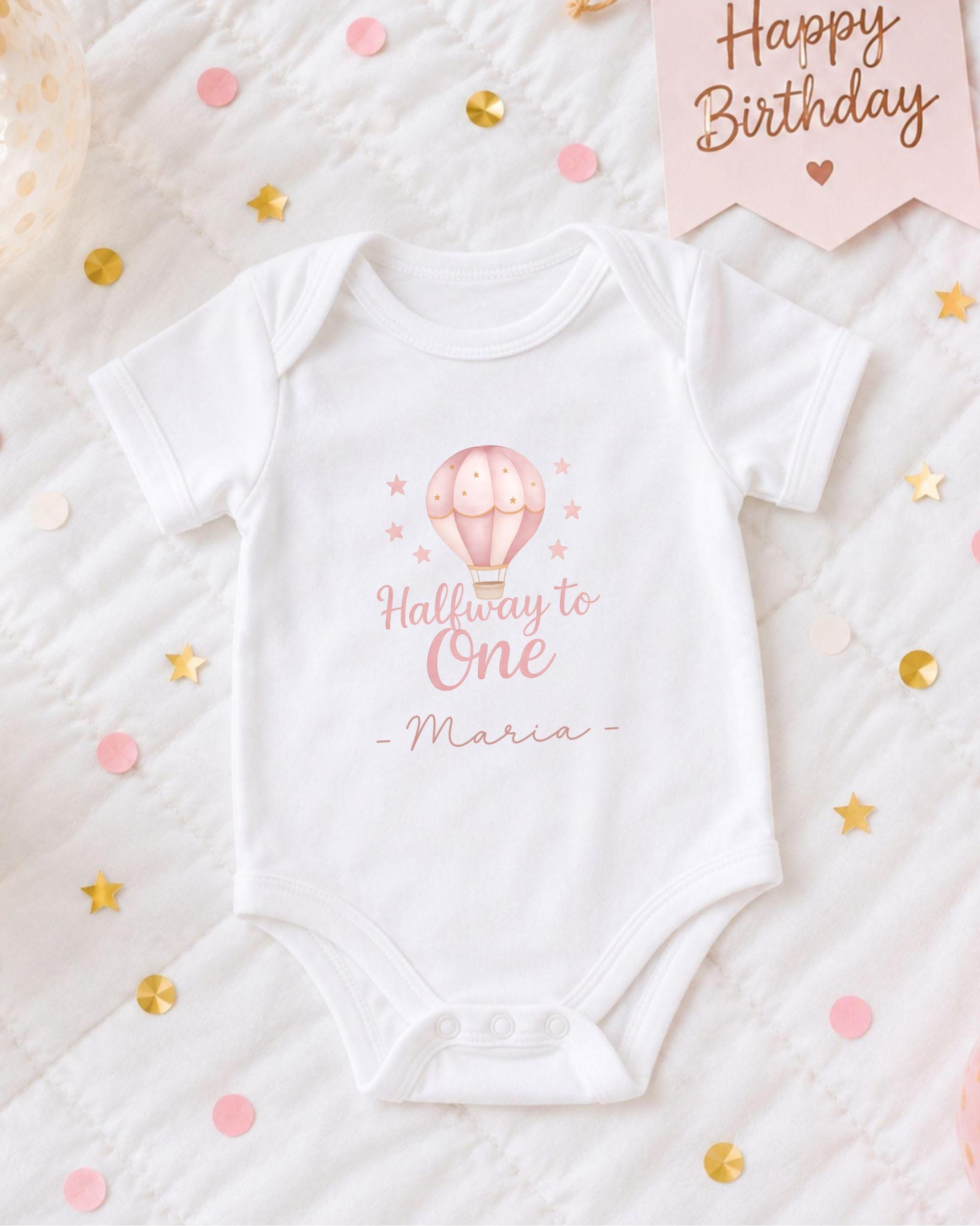 A white onesie with a hot air balloon design and the text "Halfway to One - Maria" printed on it, along with confetti and a birthday card.