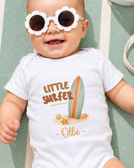 A baby wearing a white onesie with a surfboard and beach scene, smiling and wearing sunglasses.