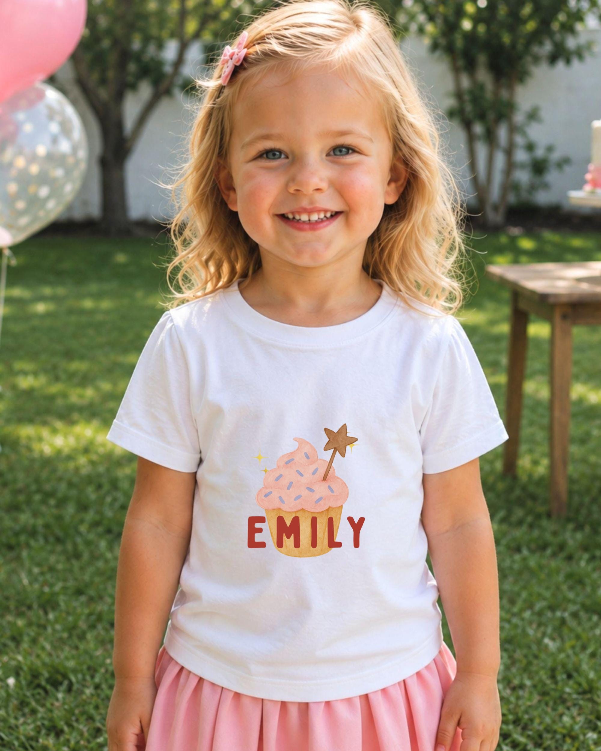 A young girl with blonde hair is smiling and wearing a white t-shirt with a cupcake design and the name "EMILY" printed on it.