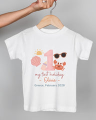 A white t-shirt with a graphic design featuring a sun, sunglasses, a shell, and a crab, along with text that says "my first holiday - Olivia - Greece, February 2028".