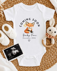 A baby announcement outfit with a fox graphic, a crocheted rabbit, and a baby announcement photo, all arranged on a woven placemat.