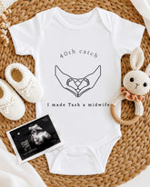 A white onesie with a graphic of hands holding baby feet, accompanied by a crocheted rabbit toy and a baby bootie. The image also includes an ultrasound image of a baby's feet.