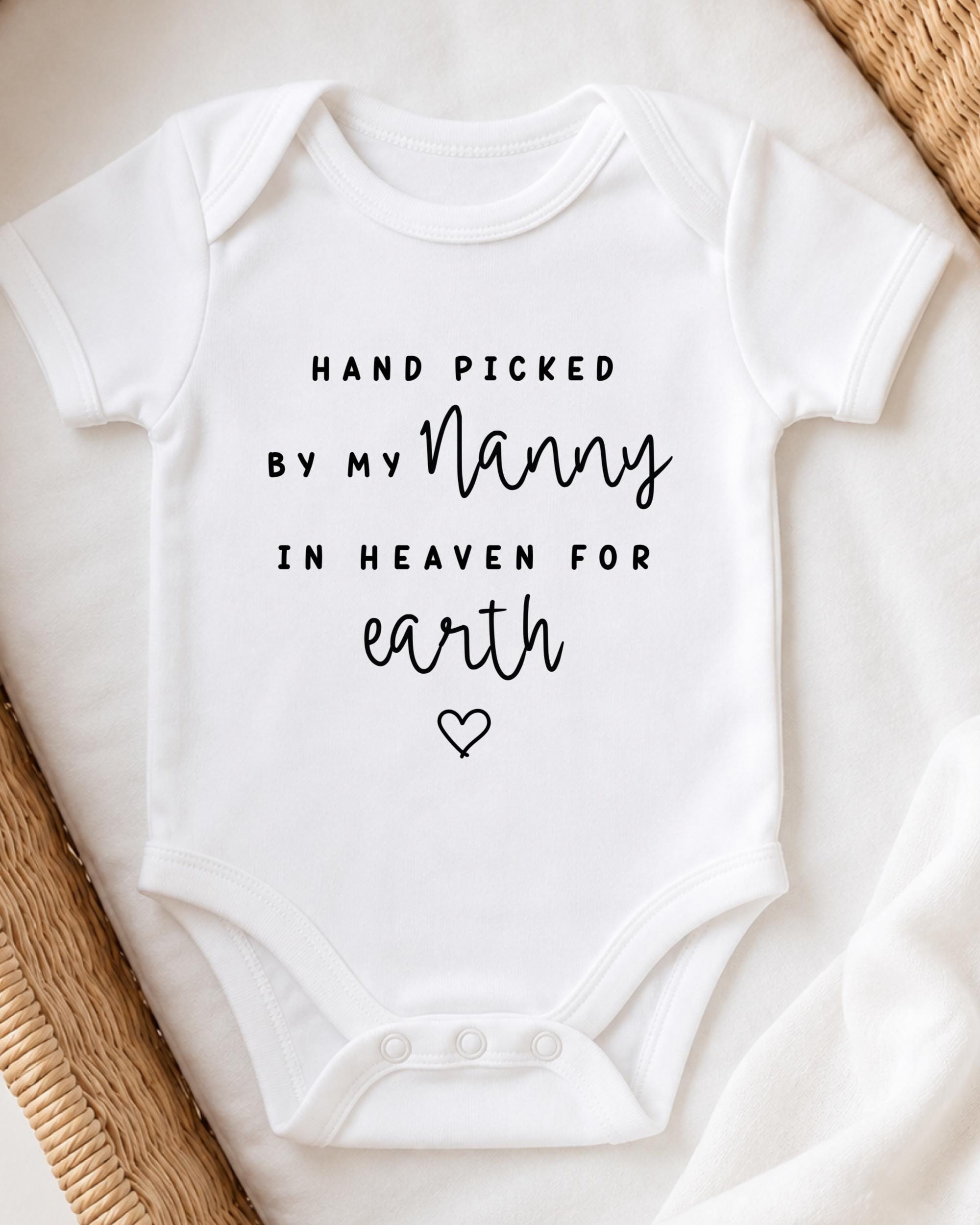 A white baby onesie with black text that reads "HAND PICKED BY MY NANNY IN HEAVEN FOR EARTH" and a heart symbol.