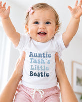A smiling baby girl wearing a white t-shirt with text and a pink bow, being held up by someone's arms.