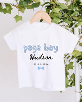 A white t-shirt with the text "Page boy Hudson" printed on it, along with a date "01.01.2022" below it. The t-shirt is hanging on a wooden hanger, and there are green leaves visible in the background.