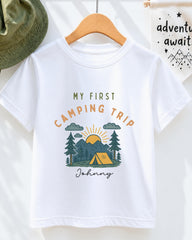 A white t-shirt with a graphic design depicting a camping scene, along with the text "My first camping trip" and "Johny" printed on it.