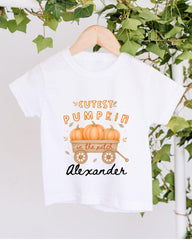 A white t-shirt with a graphic design featuring a wagon filled with pumpkins and the text "Cutest Pumpkin in the Patch Alexander" printed on it.