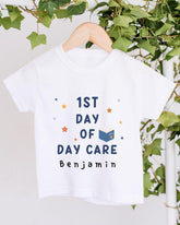 A white t-shirt with the text "1st DAY OF DAY CARE Benjamin" printed on it, along with stars and a book icon.