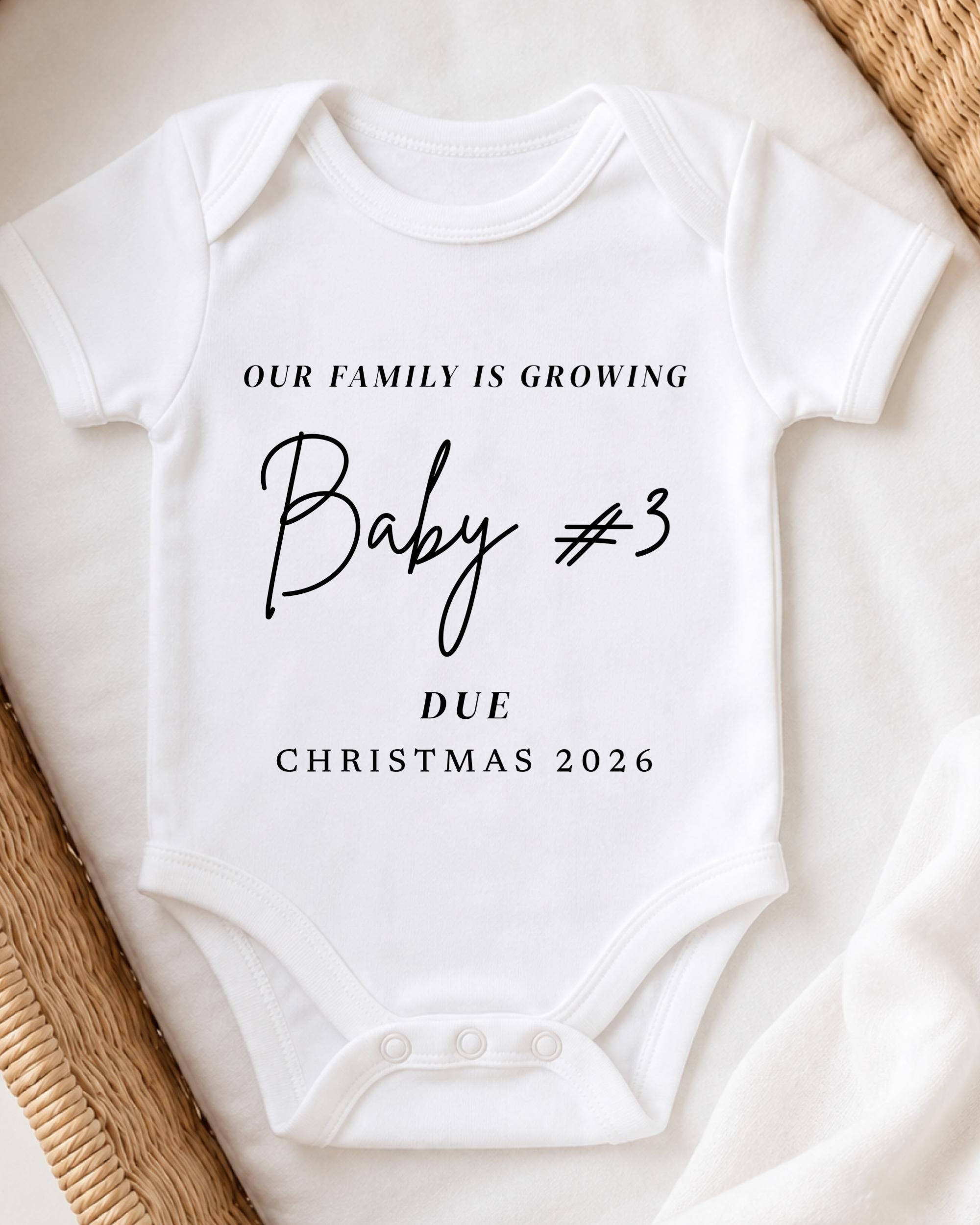 A white baby onesie with text printed on it, placed on a bed with a woven blanket.