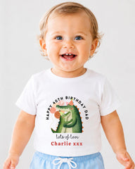 A smiling baby boy wearing a white t-shirt with a graphic of a green alligator and text that says "Happy 40th Birthday Dad lots of love Charlie xxx".