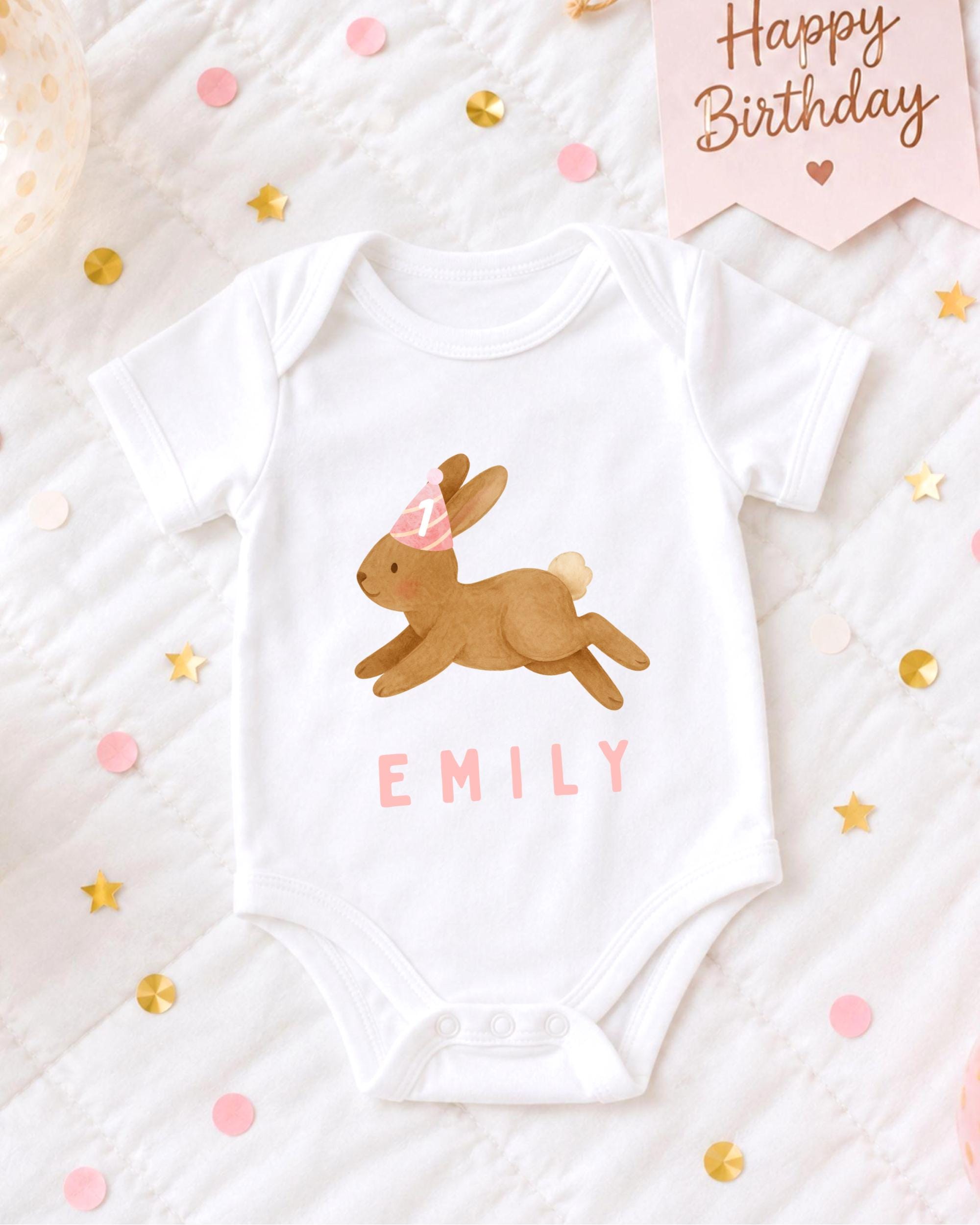 A white baby onesie with a bunny design and the name "EMILY" printed on it, along with a pink birthday banner and confetti.