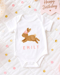 A white baby onesie with a bunny design and the name "EMILY" printed on it, along with a pink birthday banner and confetti.