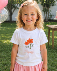 A young girl with blonde hair is smiling and wearing a white t-shirt with a flower design and the name "Poppy" printed on it.