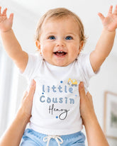 A smiling baby wearing a white shirt with the text "Little Cousin Henry" is being held up by someone's arms.
