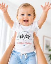 A smiling baby wearing a white t-shirt with a graphic design and text, being held up by someone's arms.