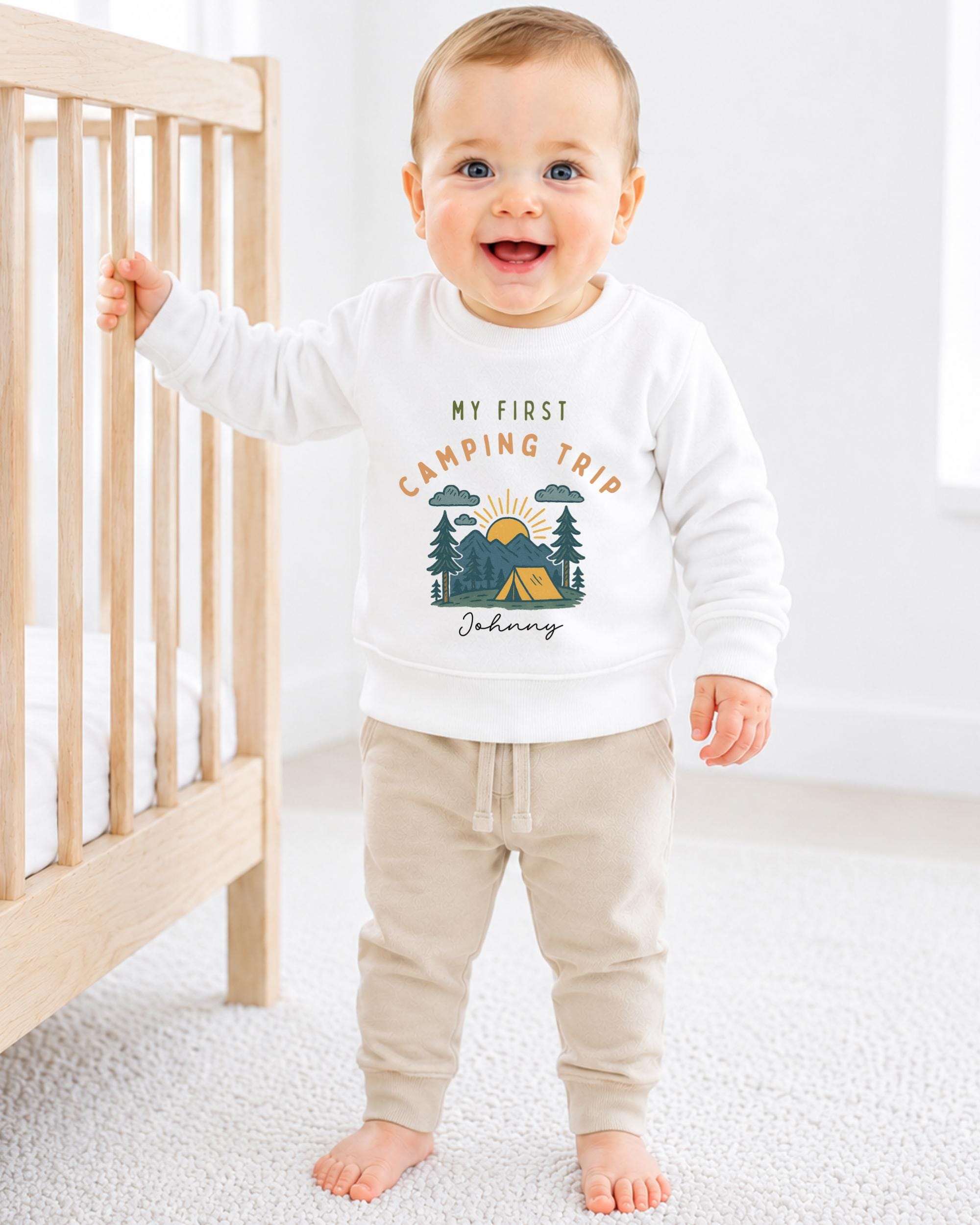 A smiling baby boy wearing a white sweatshirt with a camping trip design and the text "My first camping trip" is standing next to a wooden crib.