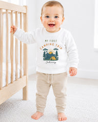 A smiling baby boy wearing a white sweatshirt with a camping trip design and the text "My first camping trip" is standing next to a wooden crib.