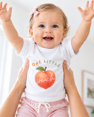 A smiling baby girl wearing a white shirt with a peach graphic and the text "One Little Peach" is being held up by someone's arms.