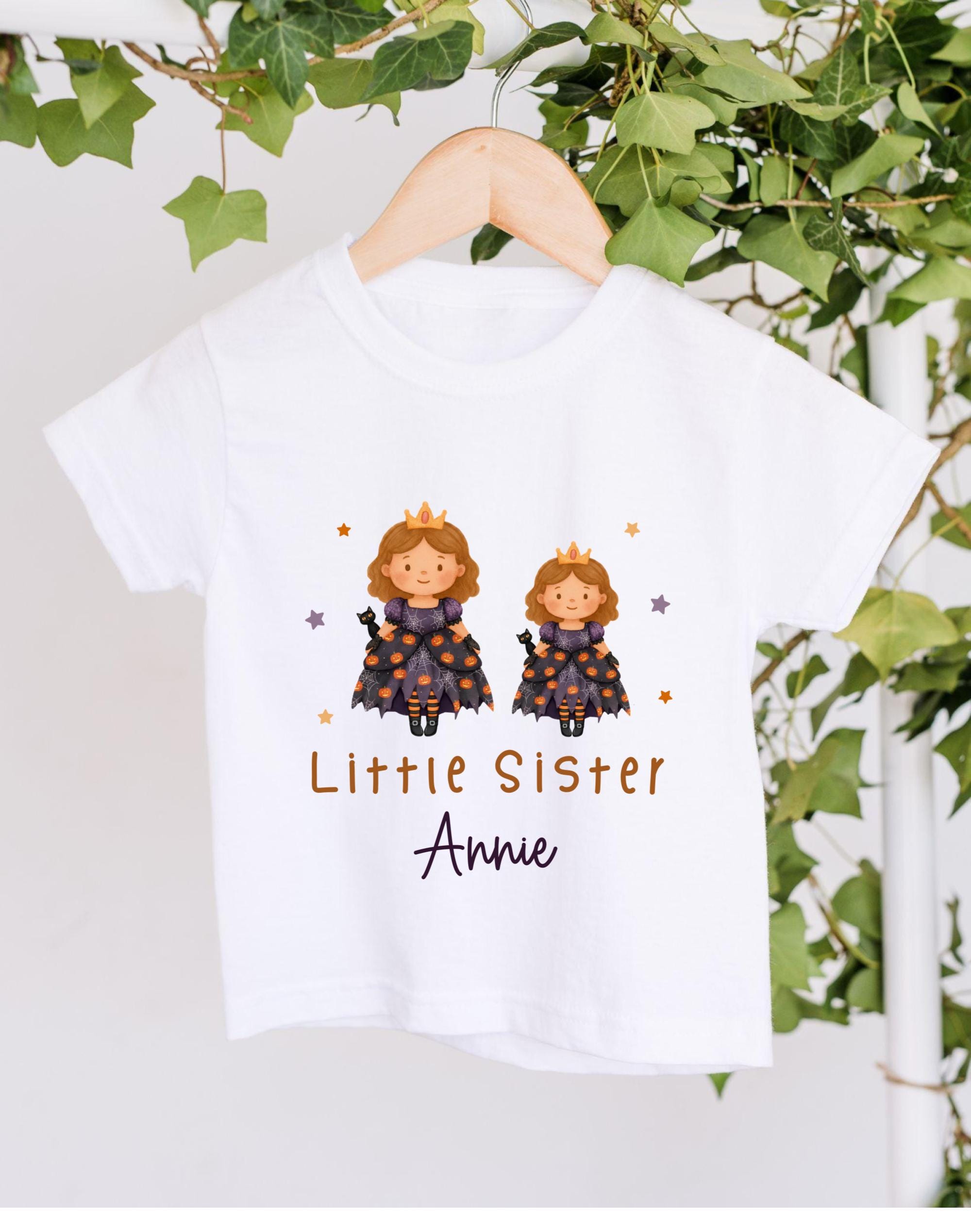 A white t-shirt with a graphic design featuring two cartoon princesses wearing crowns, surrounded by stars and vines. The text below the design says "Little Sister Annie".