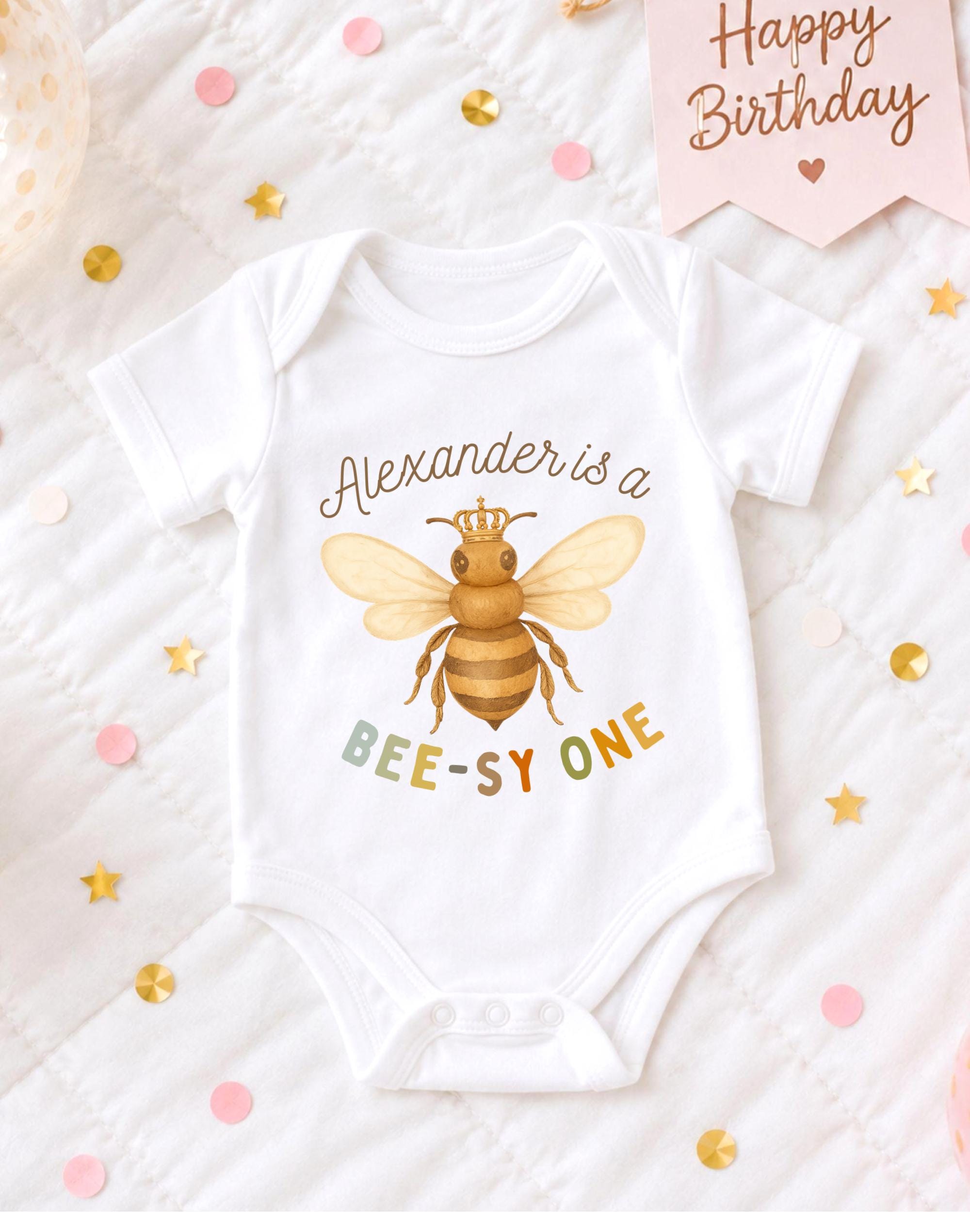 A white baby onesie with a bee design and the text "Alexander is a BEE-SY ONE" printed on it, along with confetti and a birthday card.