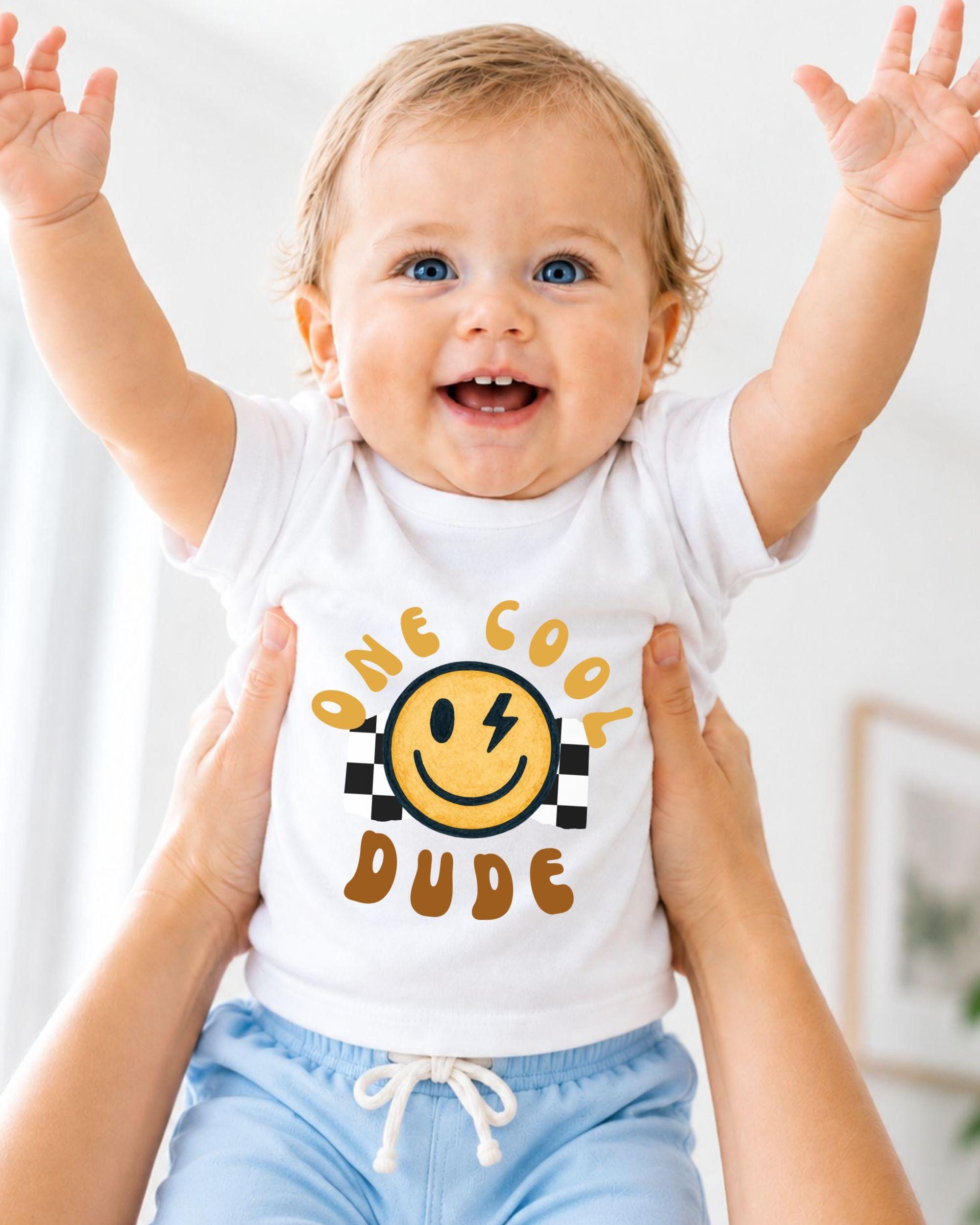 A smiling baby wearing a white t-shirt with a yellow smiley face and the text "One cool dude" is being held up by someone's arms.