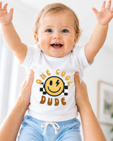 A smiling baby wearing a white t-shirt with a yellow smiley face and the text "One cool dude" is being held up by someone's arms.