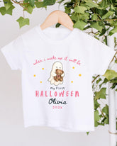 A white t-shirt with a Halloween-themed design hanging on a wooden hanger. The design includes a ghost holding a teddy bear, and text that reads "When I wake up it will be My First Halloween Olivia 2023".