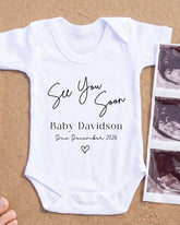 A white baby onesie with text that reads "See You Soon Baby Davidson Due December 2026" printed on it, along with a heart symbol. The onesie is placed next to a photograph of an ultrasound image.
