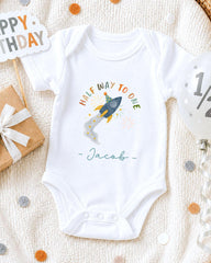 A white onesie with a rocket ship design and the text "Half Way to One" printed on it, along with the name " Jacob" written below. The onesie is placed on a white blanket, surrounded by various party decorations such as a birthday sign, a balloon, and confetti.